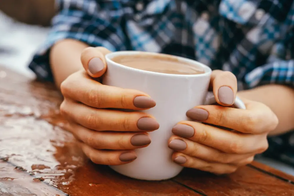 Close-up beautiful female hands hug white cup with hot drink on a wooden table outdoor