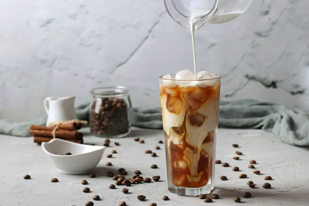 Cold coffee in a tall glass with cream being poured into it showing the texture of the drink