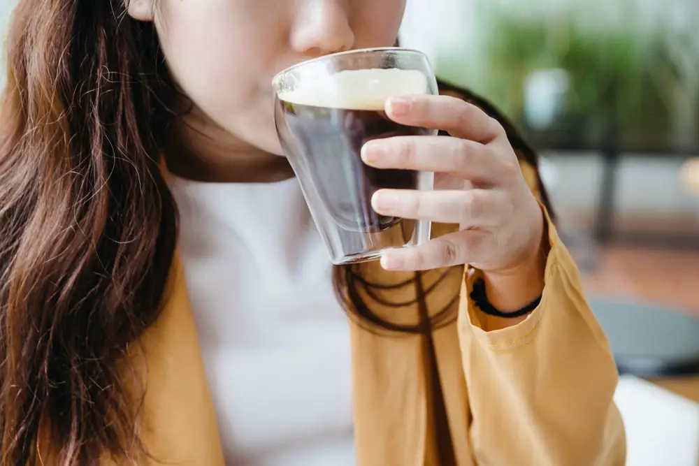 Woman wearing yellow jacket sipping Frothy Nitro Cold Brew Coffee in drinking glass with green blur background